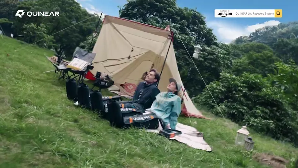 Two people relaxing in QUINEAR air compression leg recovery boots beside a camping tent in a green outdoor landscape